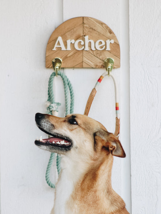 A happy dog's side profiles looks off screen to the left. Behind her is an arch shaped leash holder with a wooden mosaic pattern. The name Archer is shown in a serif font, painted off-white. The wood is a warm, light brown.