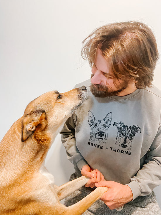 A tan colored dog reaches up to offer kisses to her human dad. Her dad is wearing a sweatshirt with a portrait of her and her brother on it. 